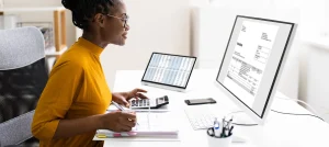 smiling young woman works with calculator and binder in front of desktop computer