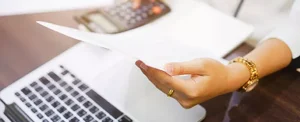 female hand holds paper in front of laptop and calculator